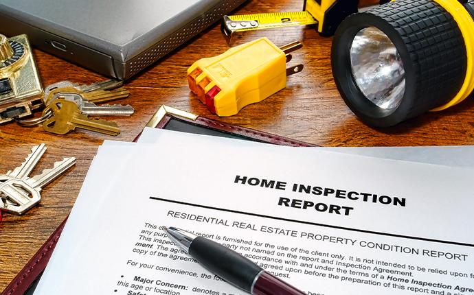 A home inspection report is on a table surrounded by various tools, like a flashlight, measuring tape, an electrical socket, keys, and a lock.