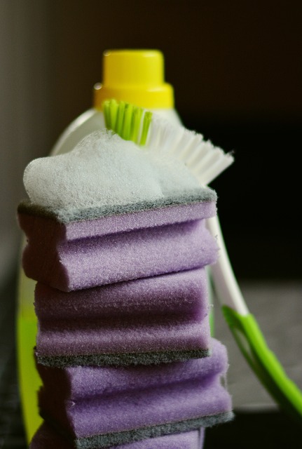 A soapy grey and purple sponge on a stack of other sponges. In the back is a cleaning brush and detergent 