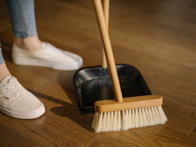 Person with light blue tapered jeans and tan shoes sweeping crumbs on a hardwood floor into a black dust pan.